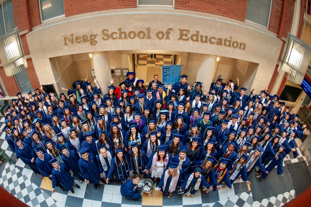 A large group of graduates in blue robes and hats pose together in the atrium of the Gentry Building at UConn Storrs. Jonathan XIV sits in front of the group.